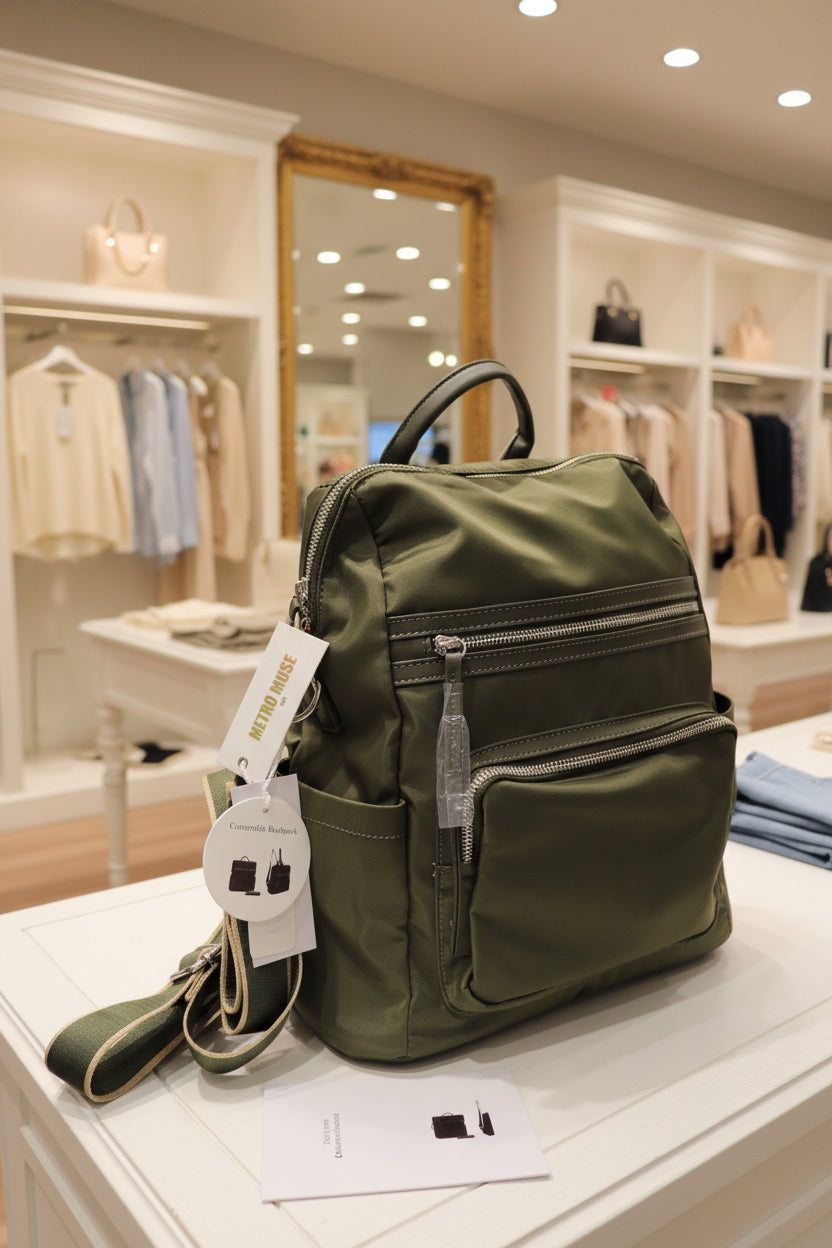 An olive green nylon backpack with multiple zippered compartments, shown with its white 'Metro Muse' brand tags attached, surrounded by plastic bags and other items on a table.