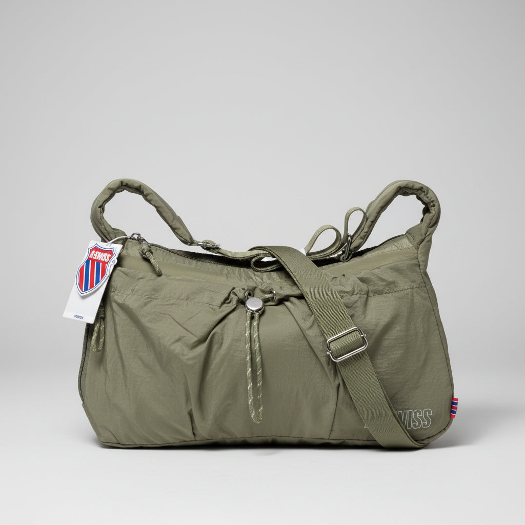 An olive green KESWISS drawstring shoulder bag with a white brand tag still attached, resting on a patterned surface with a cluttered background.