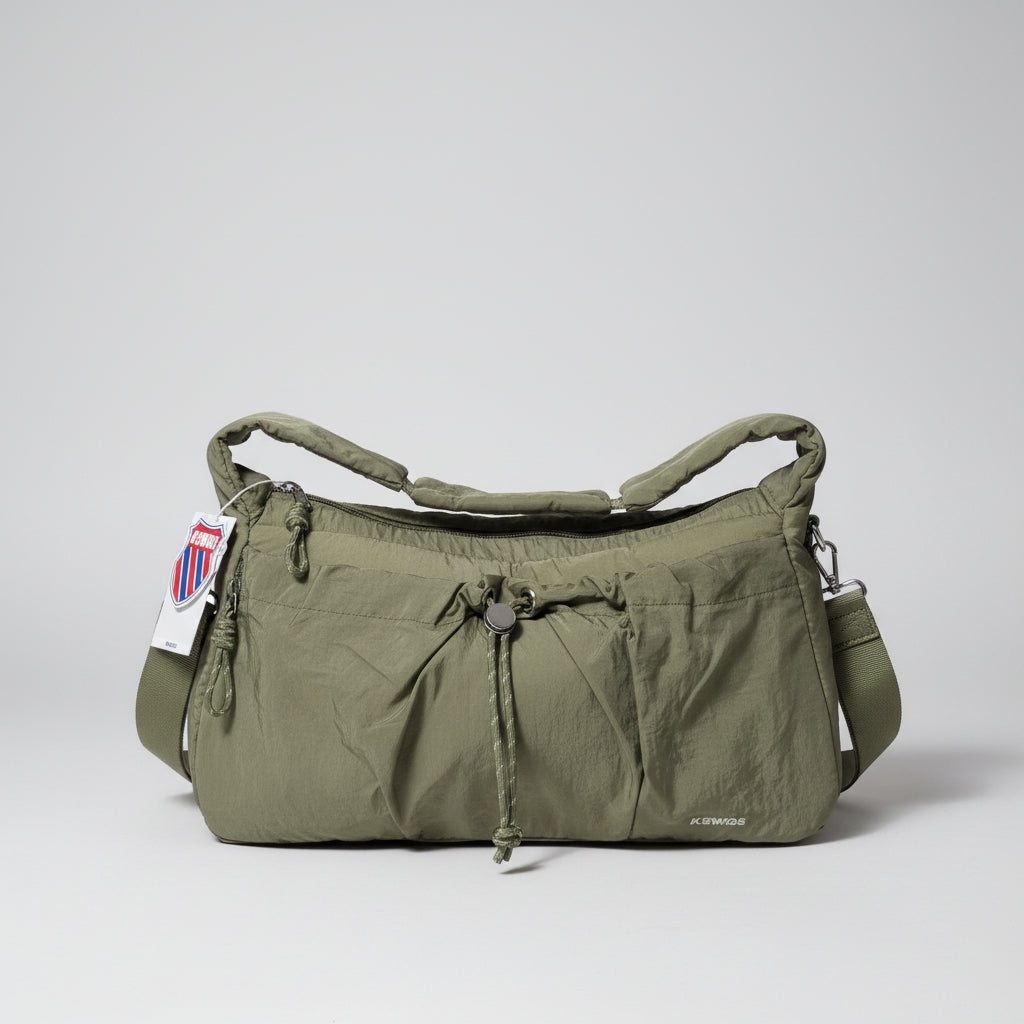 An olive green nylon shoulder bag with a drawstring closure and top zipper, shown in a cluttered indoor setting with plastic bags and boxes in the background.
