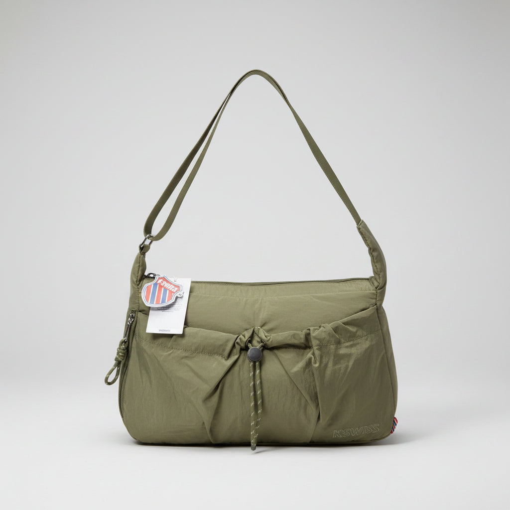 An olive green nylon shoulder bag with a drawstring closure and a hanging tag, sitting on a patterned stool surrounded by cluttered plastic bags and other items.