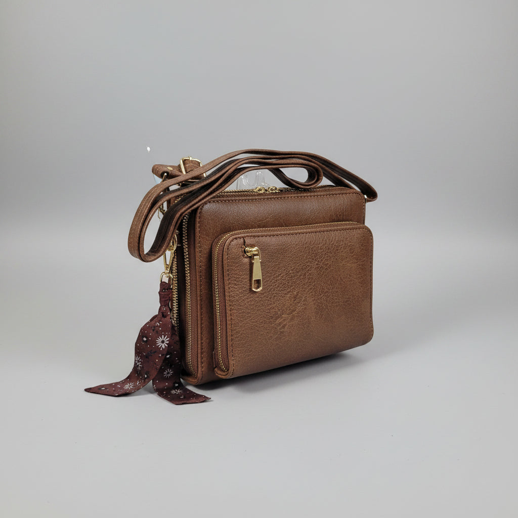 A brown faux-leather crossbody bag with multiple zippered compartments and gold-tone hardware, shown in a cluttered indoor setting.