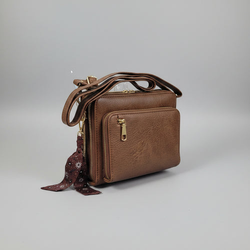 A brown faux-leather crossbody bag with multiple zippered compartments and gold-tone hardware, shown in a cluttered indoor setting.