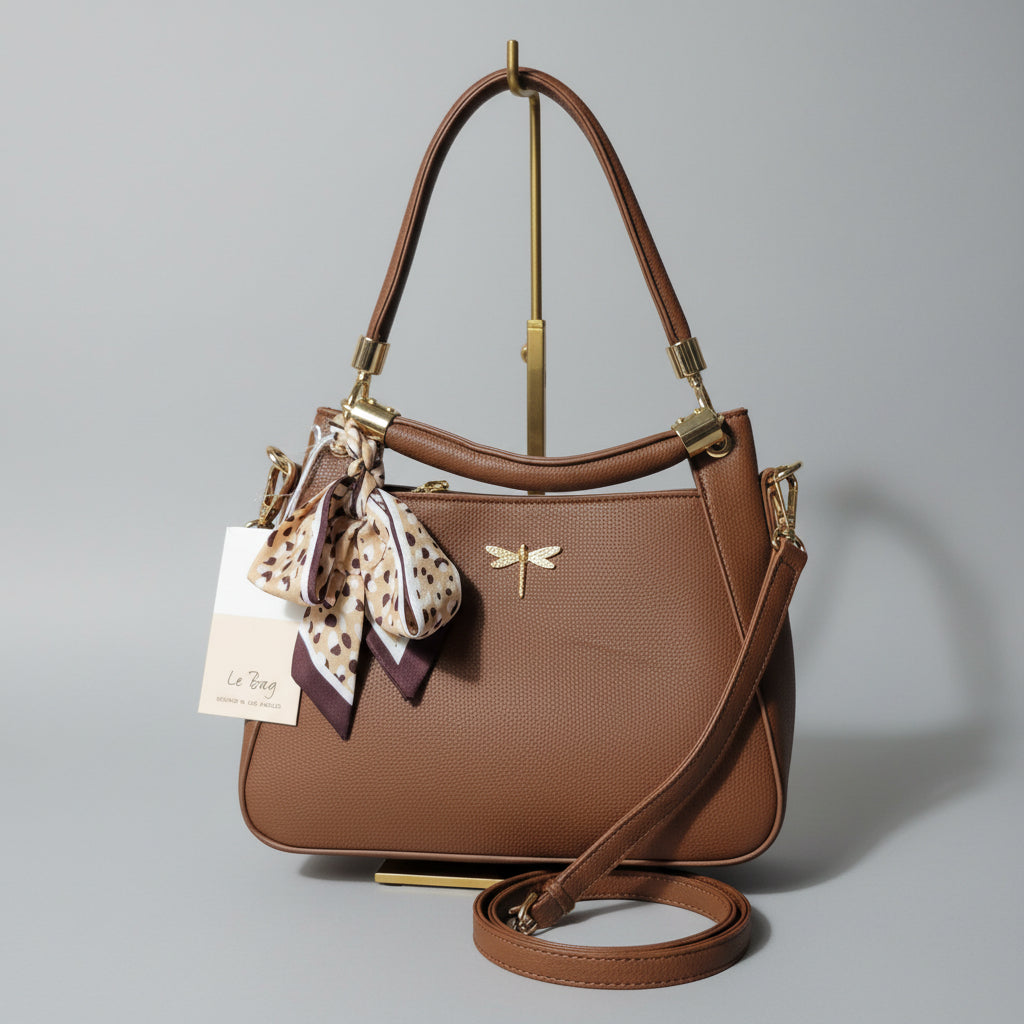 A brown leather shoulder bag with gold-tone hardware, a patterned scarf, and a white product tag, photographed in a cluttered indoor setting.